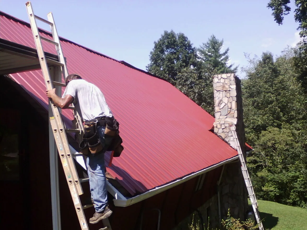 Roofer on a ladder beside a red metal roof and stone chimney — Soffit & Fascia Repair by Roofing Brothers in Mc Allister